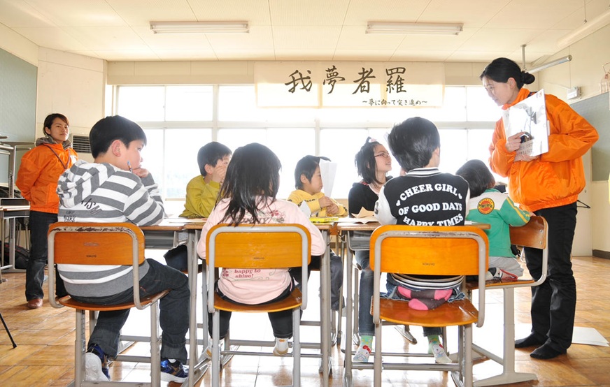 Two female teachers supervise children seated at a group of desks as they participate in a classroom activity. (Japan, 2011)