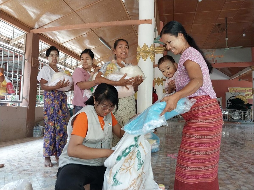A woman carrying a baby receives relief goods from a World Vision staff as other women already carrying bags of rice line up behind them. (Myanmar, 2024)