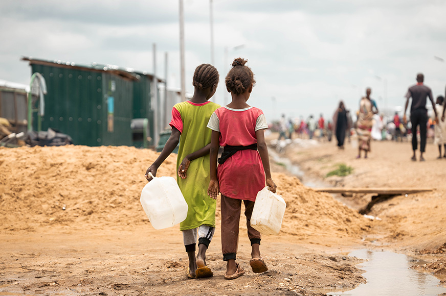 Two little girls carrying water jugs walk side by side on a dirt field beside a shallow stream.