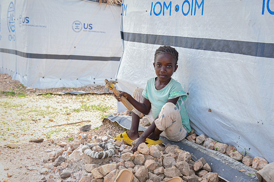 In an internally displaced persons camp, a little girl sits on the ground surrounded by rocks she’s grinding into gravel.