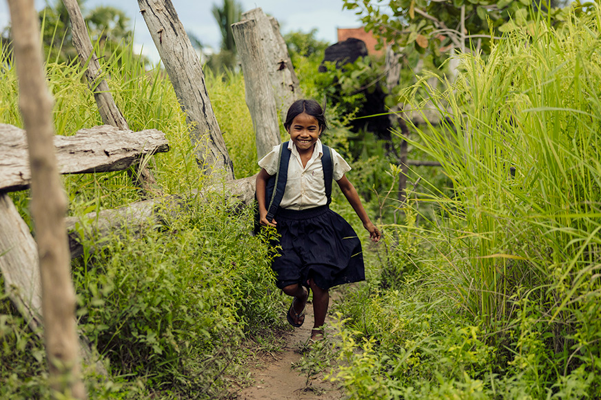 A young girl wearing a school uniform smiles as she runs on a path in a grassy field.