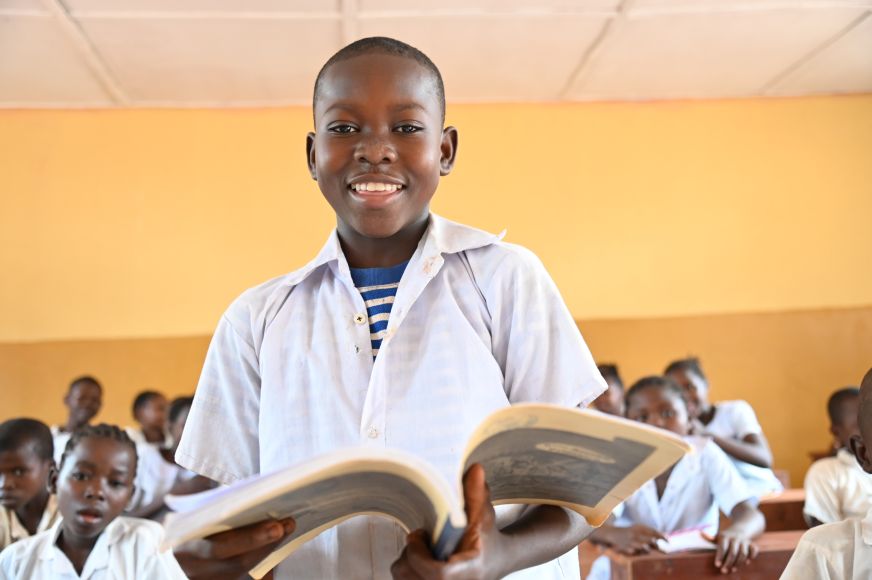 An adolescent boy in the Democratic Republic of Congo stands smiling in a yellow-walled classroom, holding an open book.