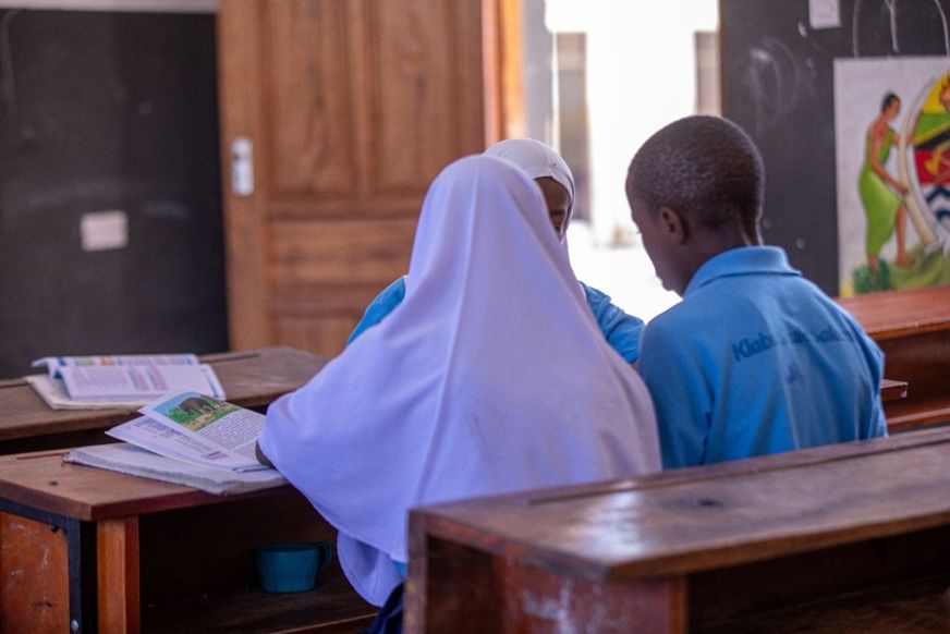 Three students, one wearing a purple hijab, sit at wooden desks in a classroom in Dar es Salaam, Tanzania.