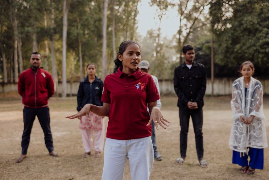 In Nepal, a young woman wearing a red polo shirt stands gesturing among a group of people.