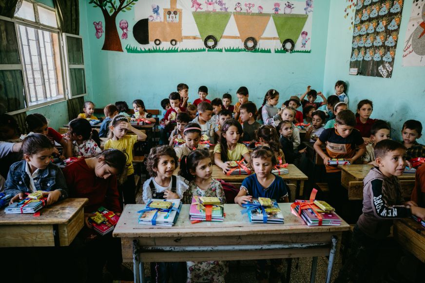 Young students crowd a colourful classroom in northwest Syria. Each learner has a package of books and supplies on the desks in front of them.