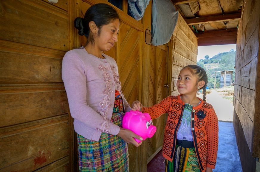 A mother in Guatemala holds a bright pink piggy bank while her daughter puts money into it.