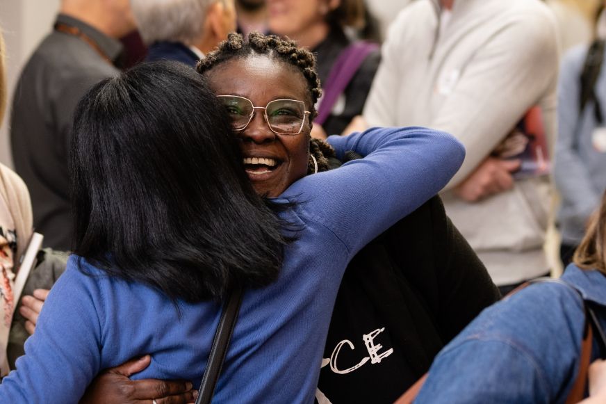 Two women joyfully embrace in a busy office environment.