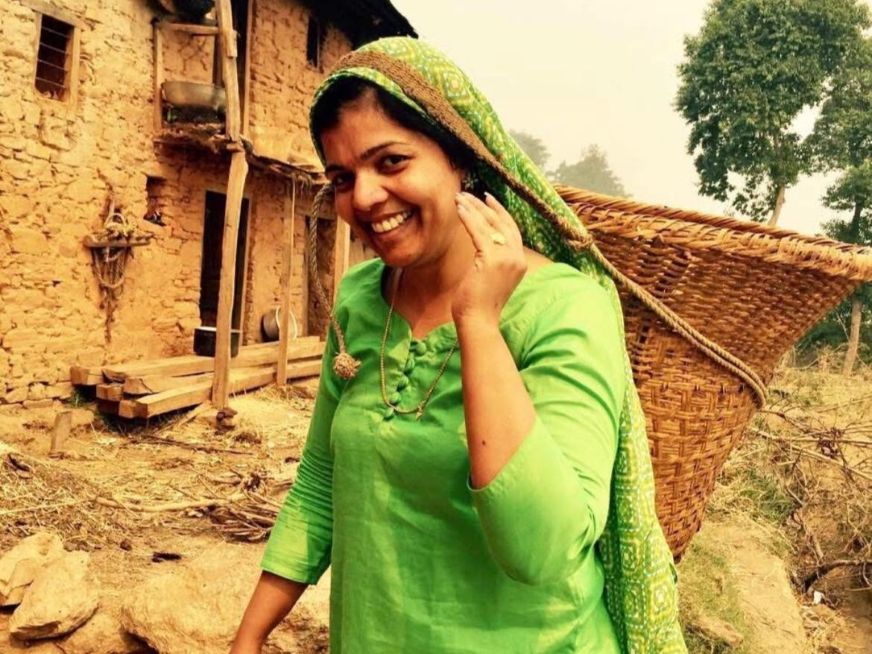 A woman wearing a green top and scarf smiles while carrying a traditional Nepali basket on her head.