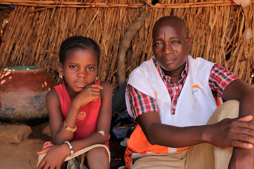 In Mali, a young girl in a red shirt sits beside a man wearing a World Vision vest.
