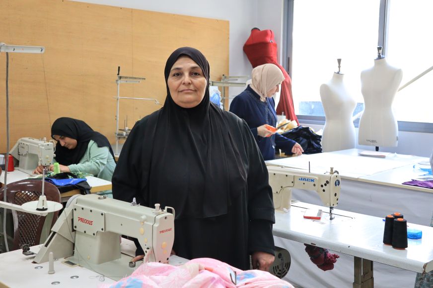 A woman in a black hijab sits behind a sewing machine in a sewing studio where other women are working.