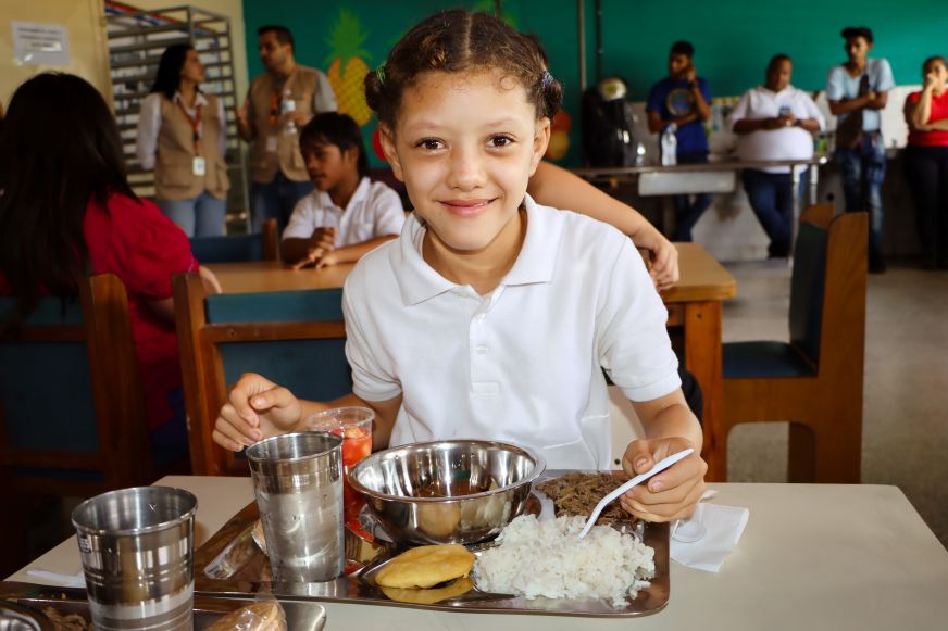 A young girl in Venezuela sits eating from a tray of food that includes rice and an arepa.