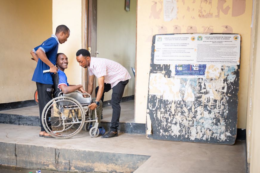 Two men help and AHADI adolescent participant in a wheelchair up some stairs.