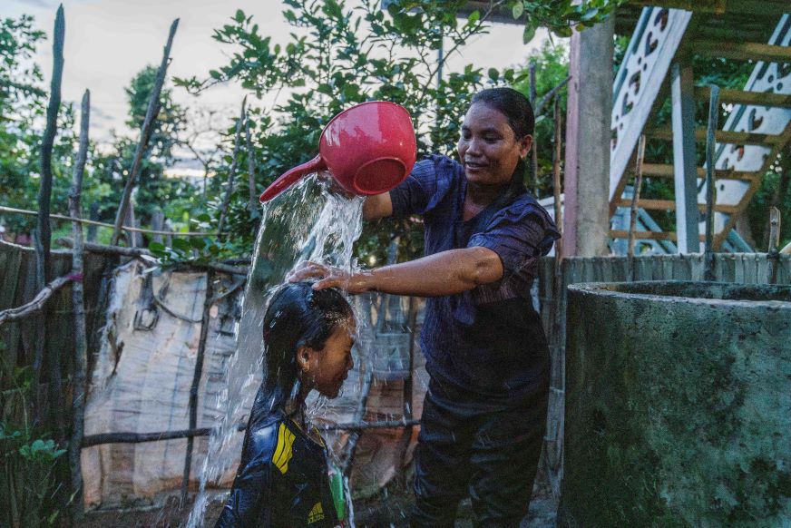 In Cambodia, a mom pours water from a red bowl onto her daughter’s head.