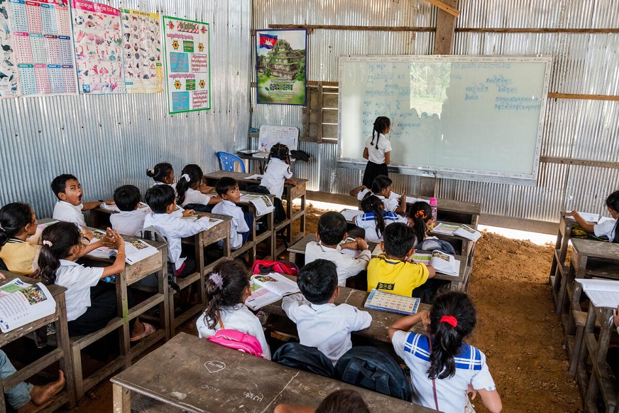 Students face a whiteboard in a classroom surrounded by wooden desks, a dirt floor and walls of corrugated steel. (Cambodia, 2024)