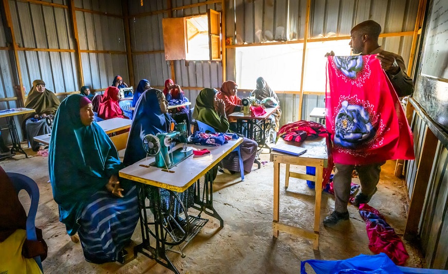 A classroom of female students sitting at desks with sewing machines, listening to their male tailoring instructor. (Somalia, 2023)