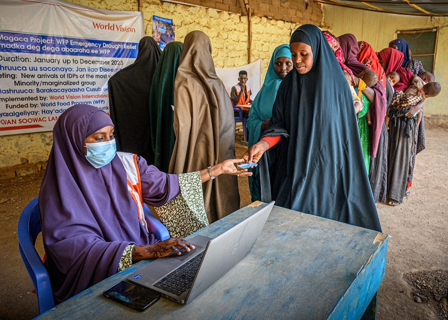 A seated woman wearing a face mask and long purple headscarf uses a laptop while reaching for a card from a woman wearing a long black headscarf and shawl. Ther