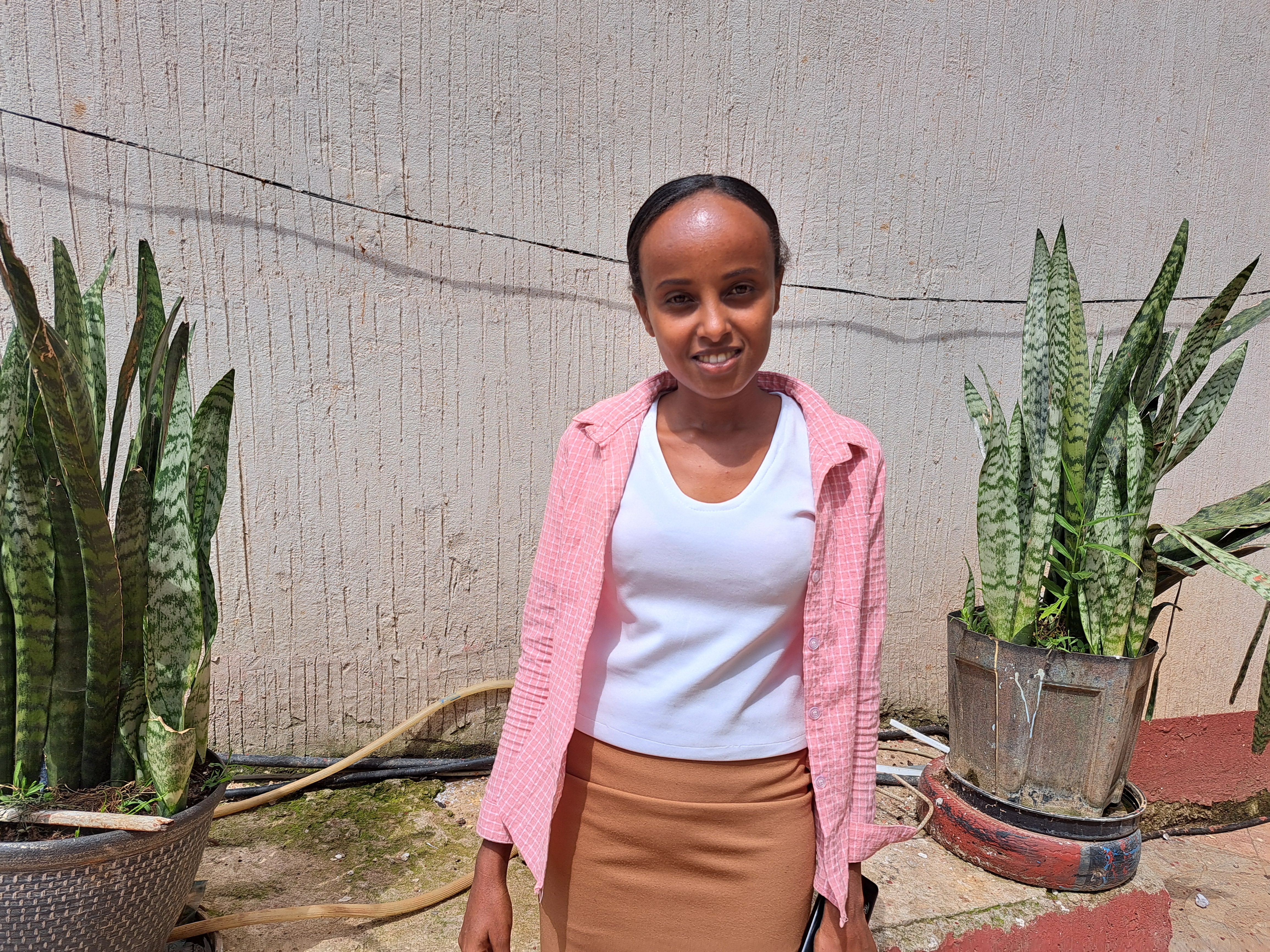 A young woman in a pink and white shirt stands in front of two potted plants and smiles.