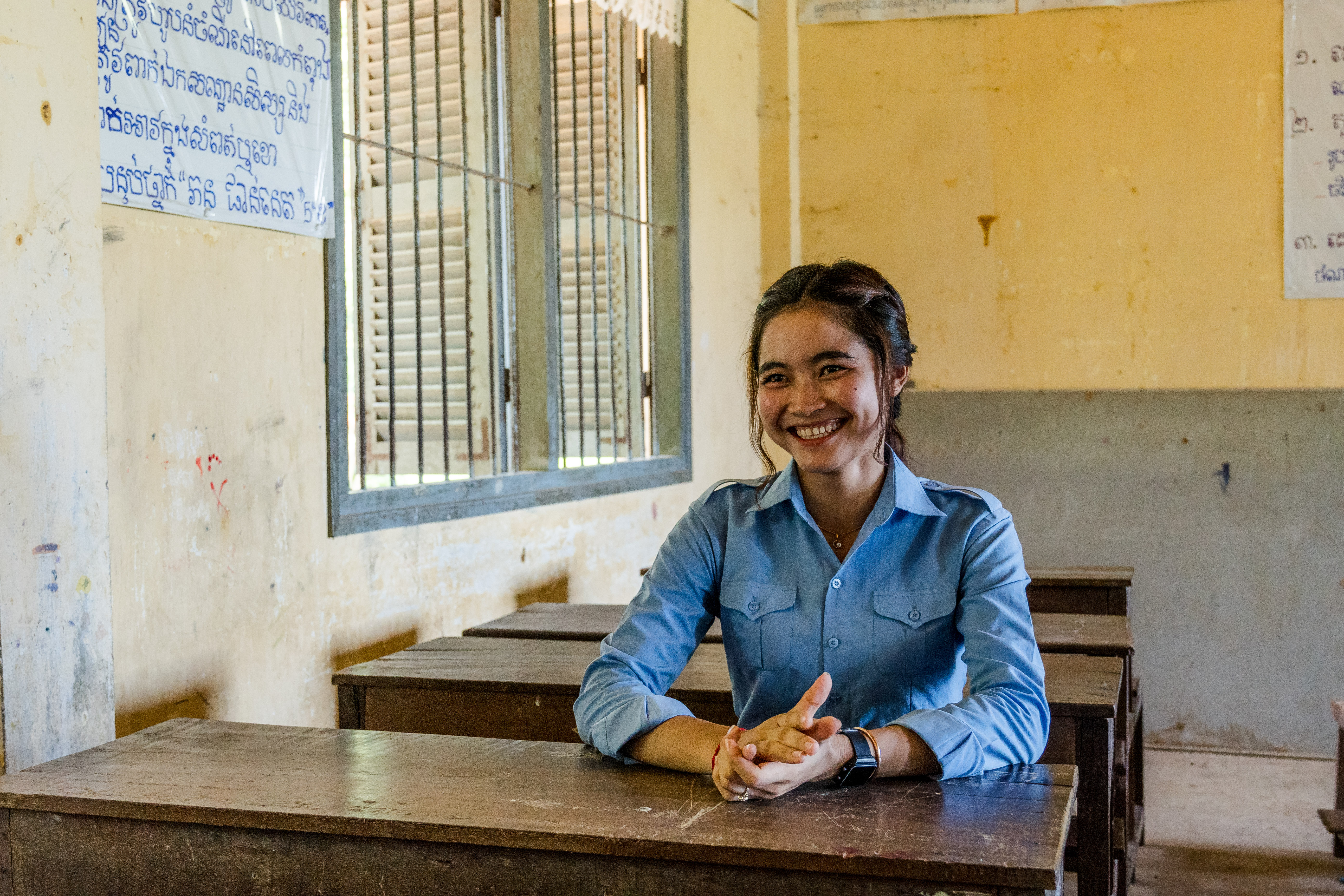 A young woman wearing a blue blouse, sits at a desk in a classroom.