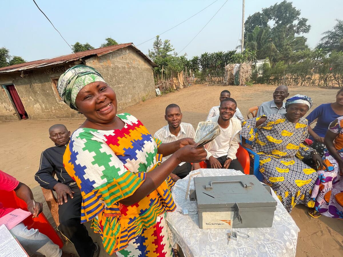 A woman in brightly coloured clothing stands in front of a lock box holding a handful of paper money, while a group of people smile in the background.