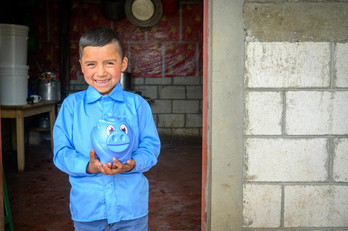 In Guatemala, a boy wearing blue smiles in a doorway holding a blue piggy bank in his hands.