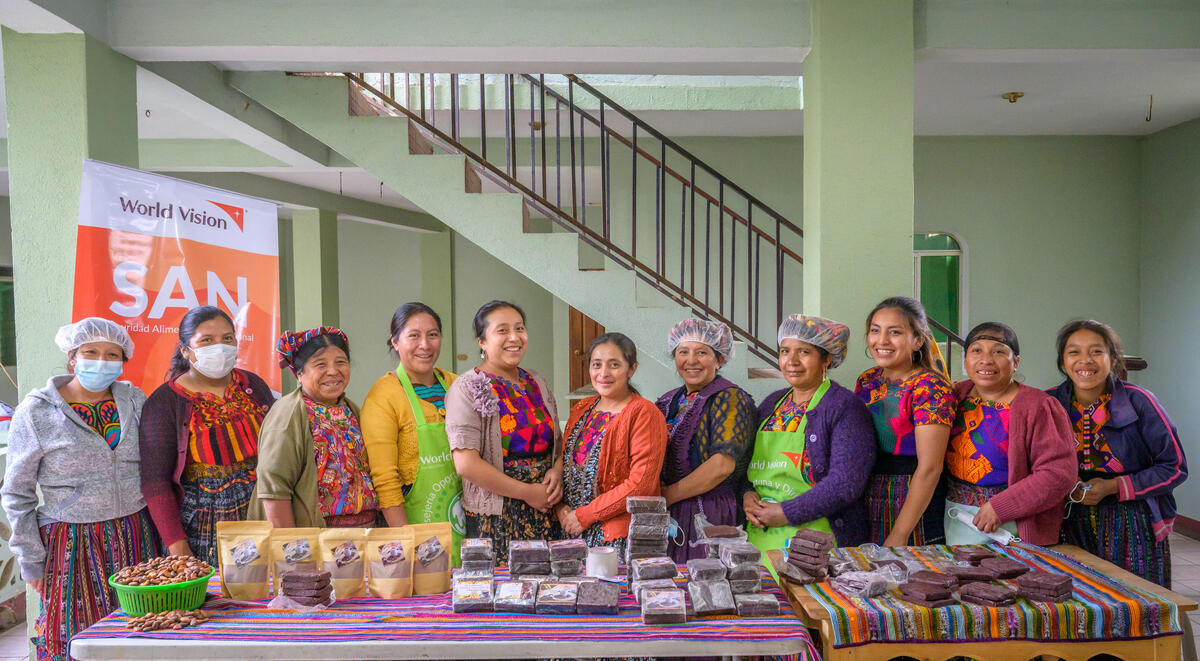 A group of women in Guatemala wear aprons and bright colours and stand behind a table covered in chocolate products.