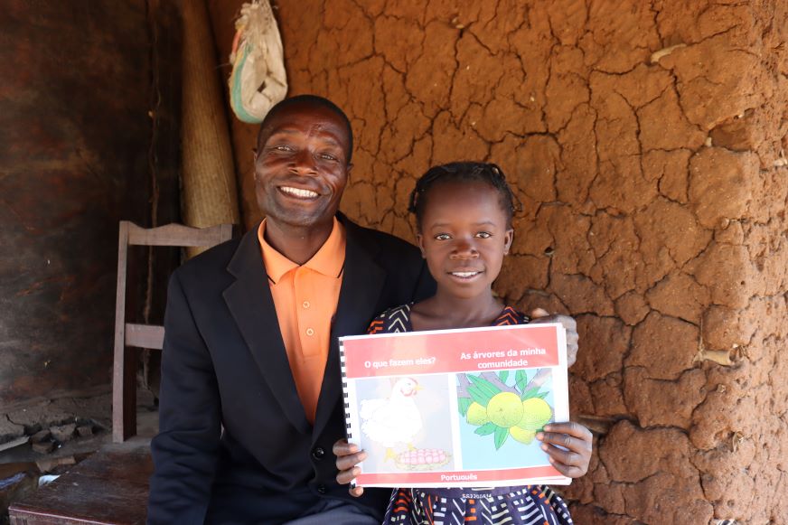 In Mozambique, a young girl holds up a book and smiles with her father.