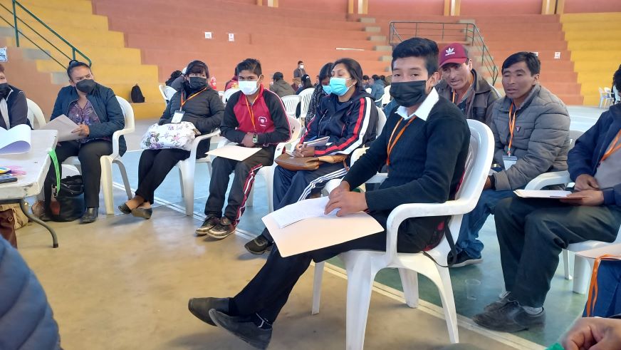 A group of teens and adults sit in white plastic chairs. Several are wearing medical masks.