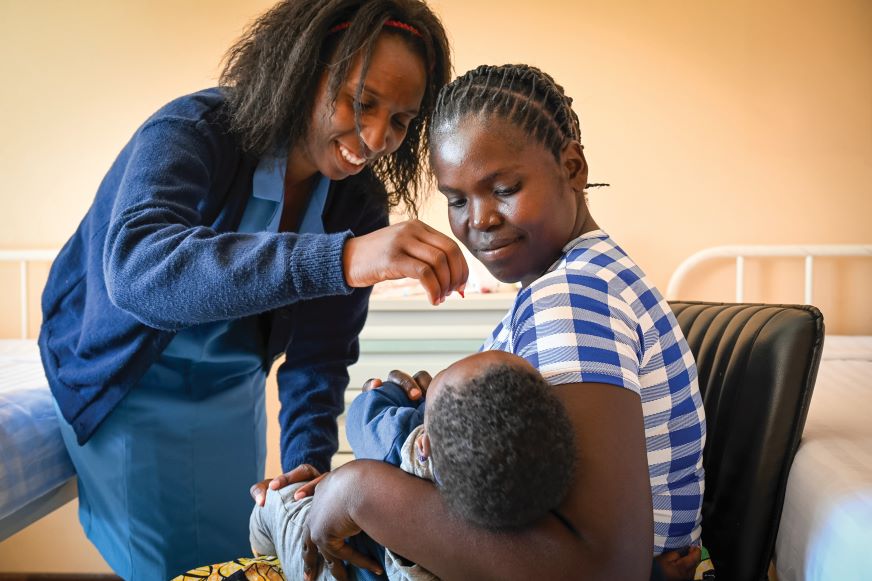 A woman holds her baby while a health worker administers a vitamin drop.