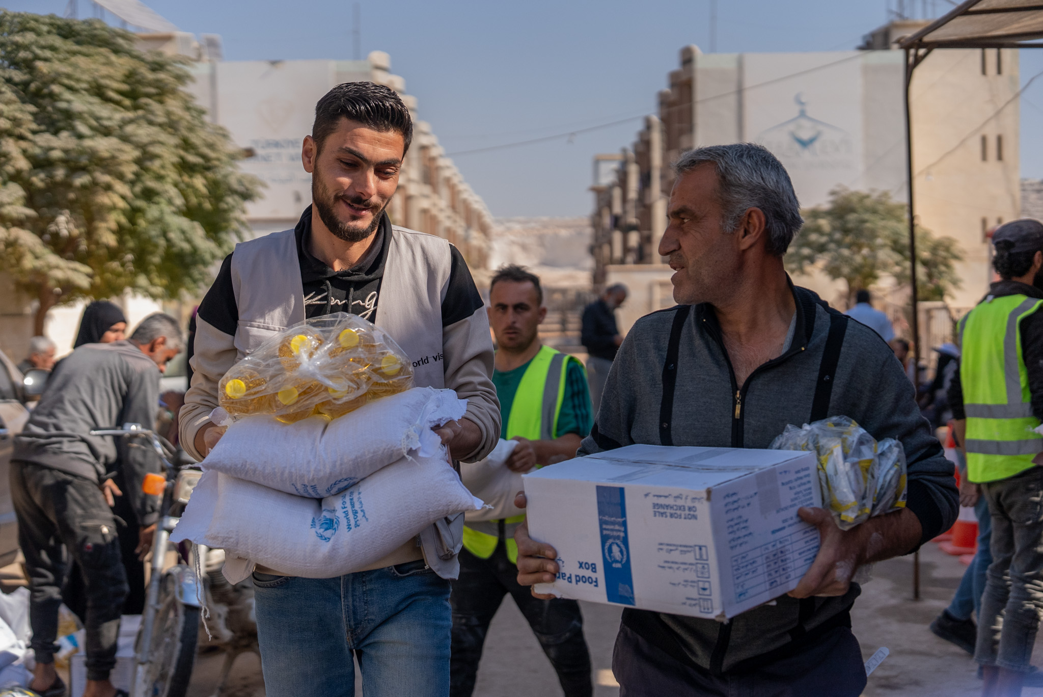 A man in a World Vision vest walks beside a father in Syria. Both carry boxes and food supplies with World Food Programme logos. (Syria 2024)