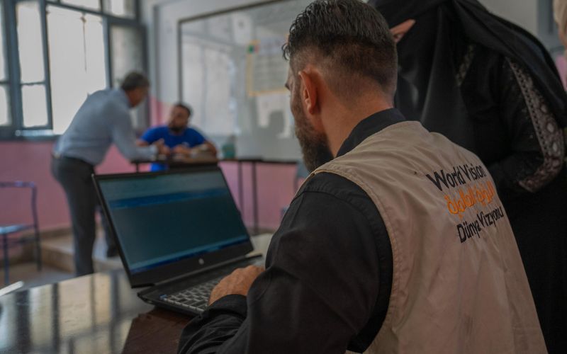 A World Vision worker types on a computer while a woman looks on at the screen, awaiting her emergency cash assistance.