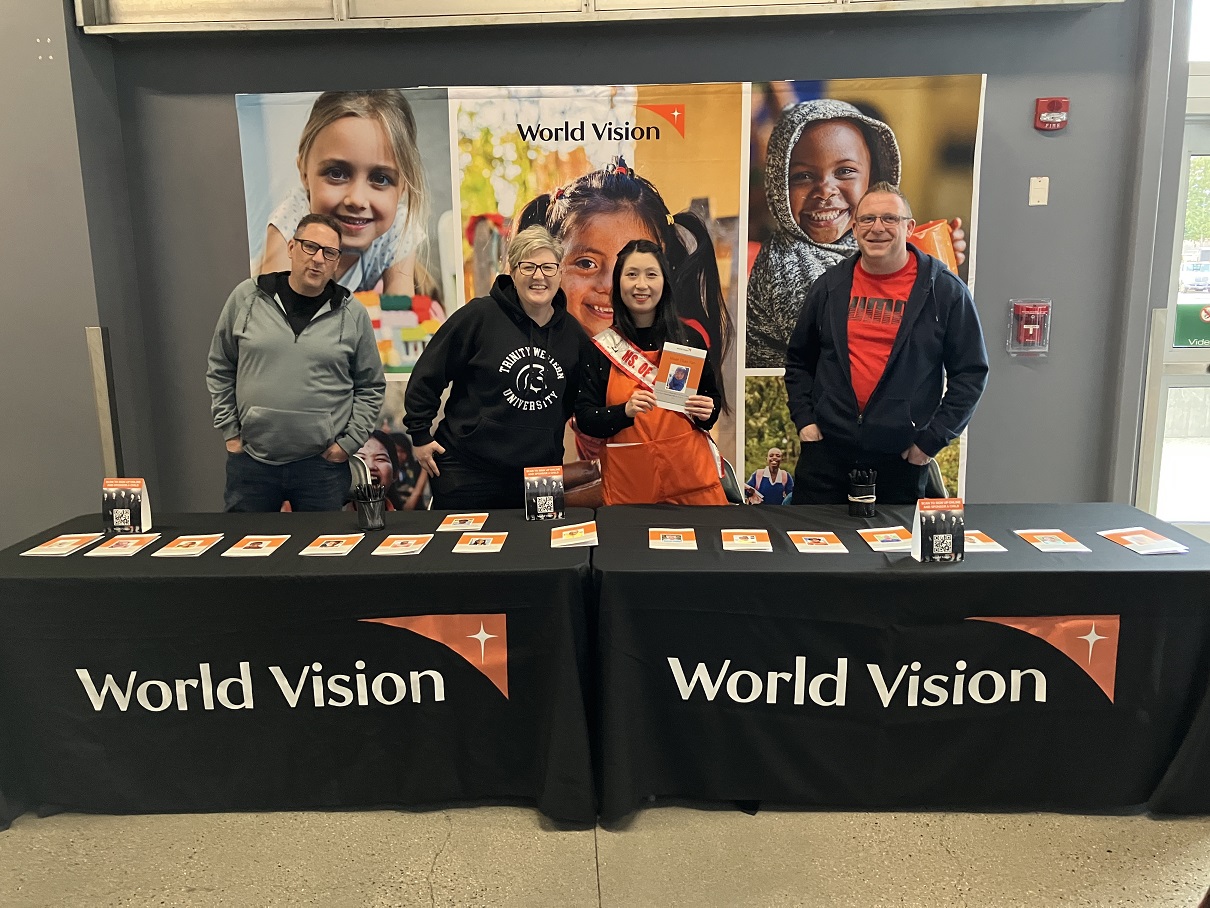 Four volunteers stand behind tables branded with large World Vision logos.