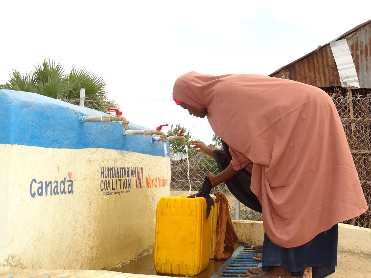 In Somalia, a woman wearing a light red hijab fills a yellow water can from a tap branded with the Government of Canada, Humanitarian Coalition and World Vision