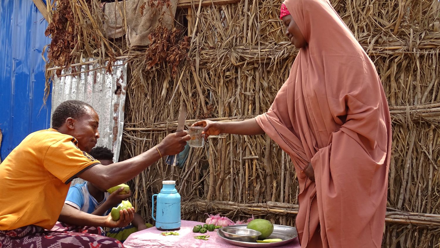 A Somali woman offers a clean glass of water to a seated man.