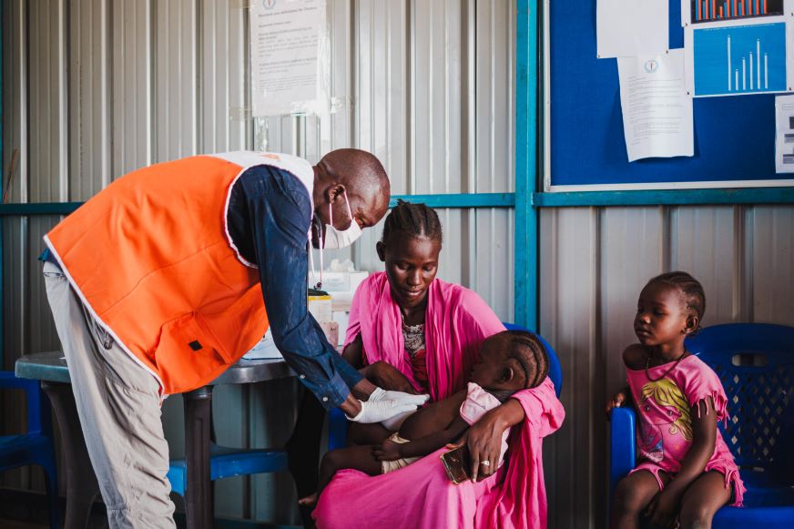 A health worker in an orange World Vision vest leans over a child in her mother's lap, while another smile child watches on.