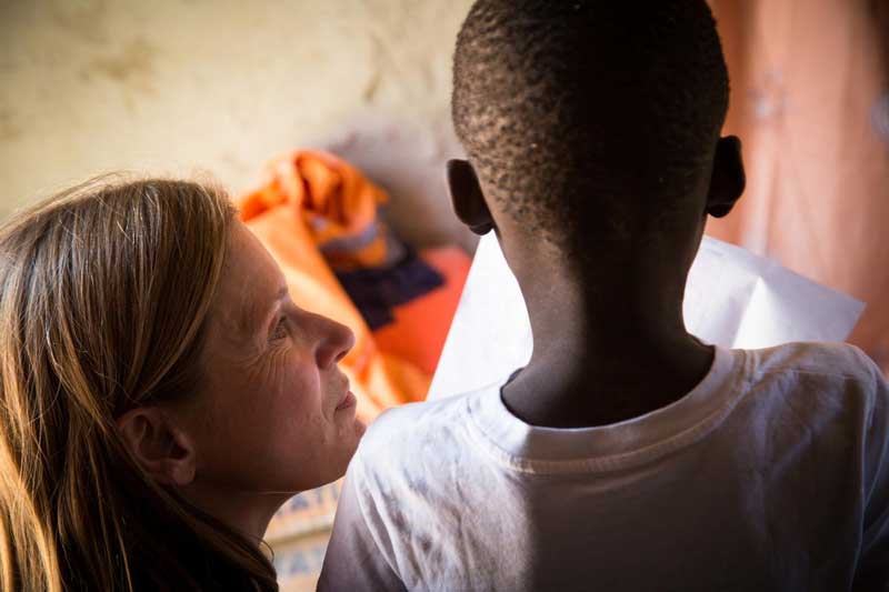 A woman kneels beside a child who is reading to her from a paper.