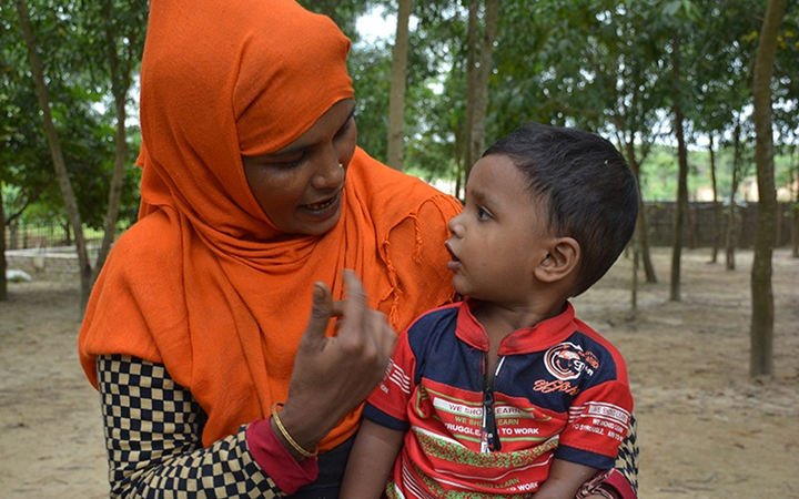 A small boy sits on the lap of a woman wearing a bright orange headscarf, looking toward her face as she speaks to him.