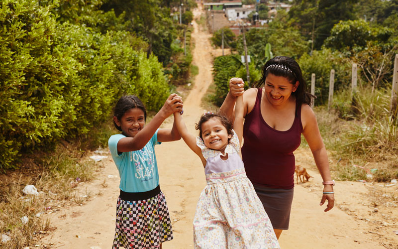 A woman and two children laugh and hold hands walking down a dirt road