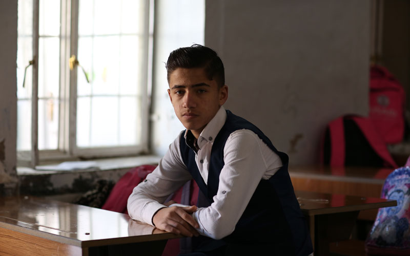 A young Iraqi boy sits at a desk and looks directly at the camera
