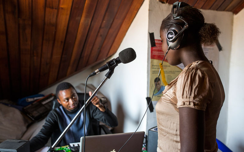 A young Congolese girl stands at a microphone while a man at a computer gives her a thumbs up sign