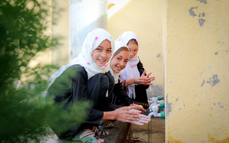 Three Afghan girls wearing white head scarves smile at the camera as they crouch down to wash their hands at a row of fawcetts