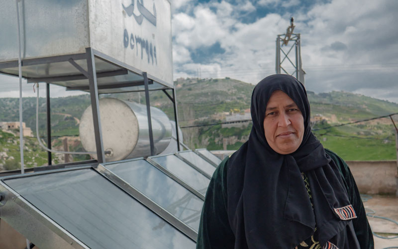 A Syrian woman stands in front of a passive solar water heater on the roof of her home in Jordan