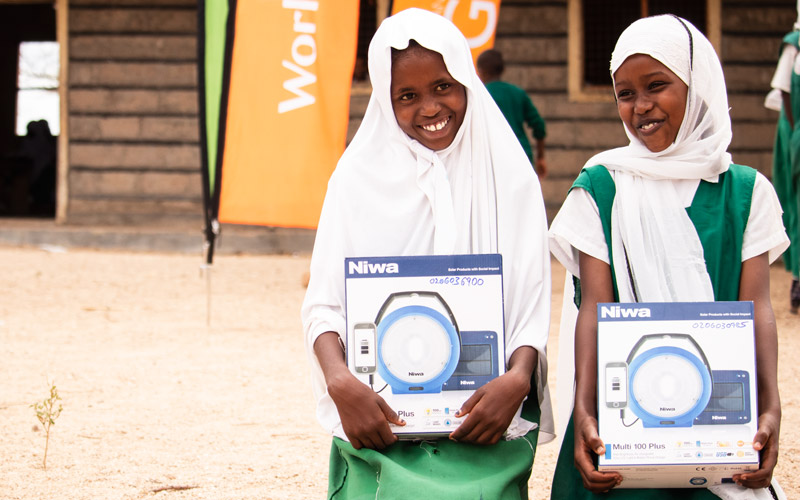 Two Kenyan girls kneel on the grown outside smiling and each holding a box with a picture of a light on it.