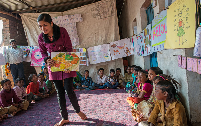 An Indian girl holds a book open to a group of children gathered in a circle.