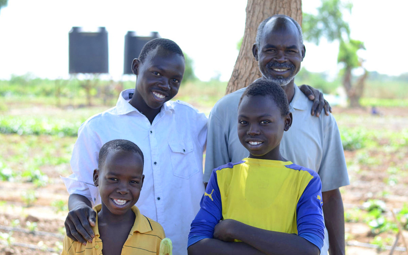A family from Sudan - dad and three young sons - stand in the shade of tree. There is garden field with water tanks in the background.
