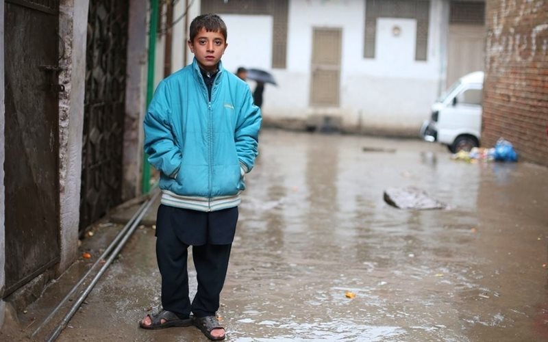 Pakistani boy wearing a blue jacket stands in an alley staring at the camera.
