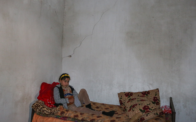 A young girl sits on a bed in a concrete room.