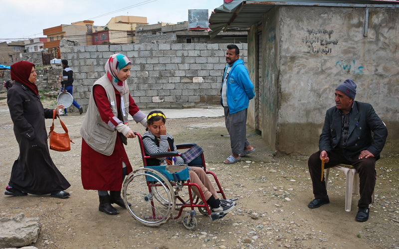 A woman pushes a young girl in a wheelchair through a yard.
