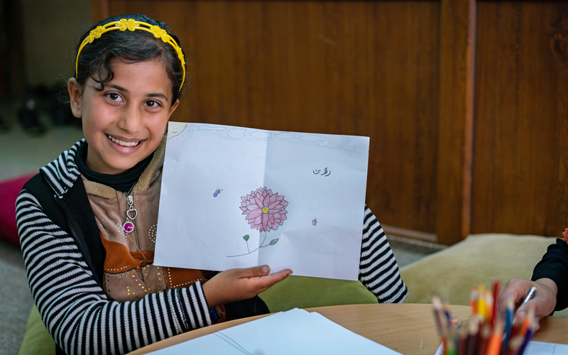 A young girl looks directly at the camera and smiles holding a piece of paper with a hand-drawn picture of a flower on it.