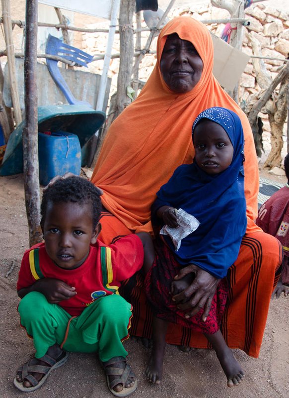 A Somali woman sits with two toddlers. The girl child holds a white package of ready-to-use-therapeutic-food (RUTF).
