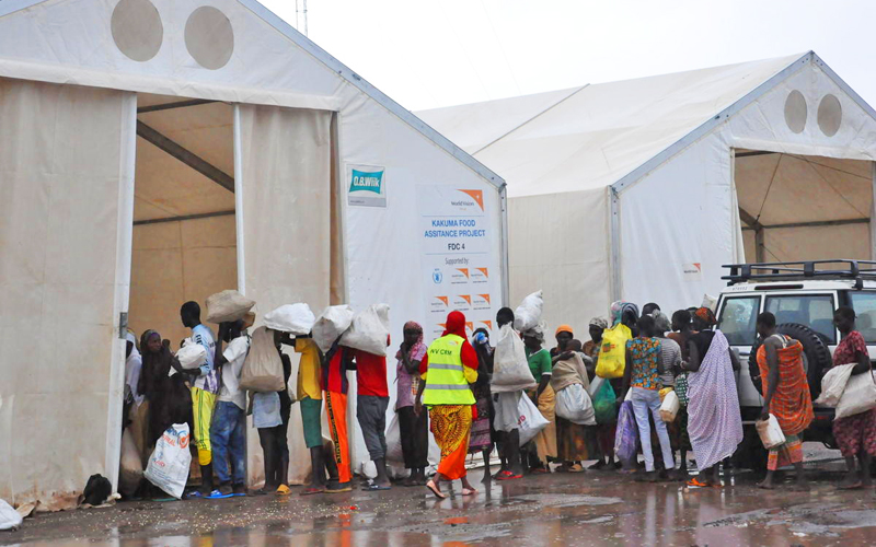 A crowd of people holding white sacks, lined up in front of two large white tents and a white SUV.